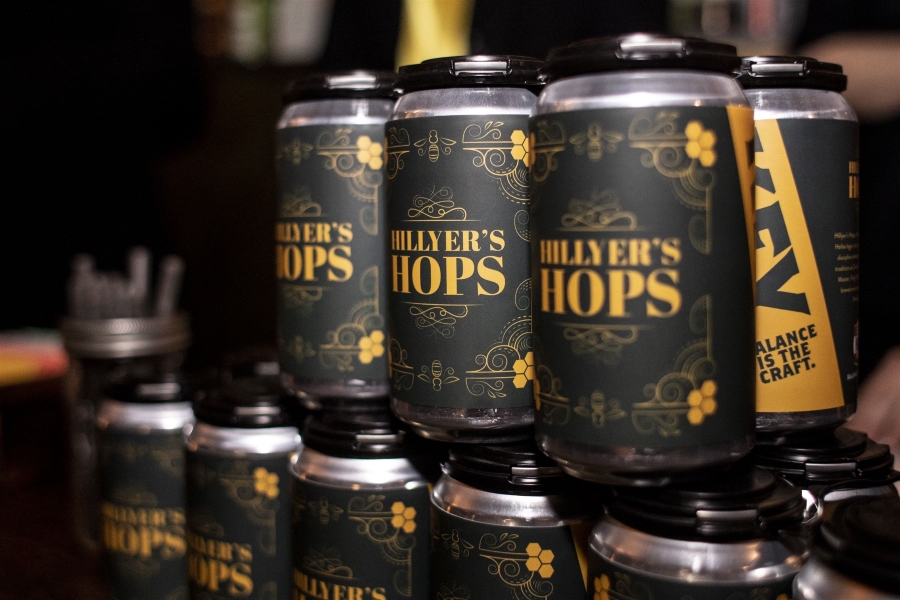 Stacked up beer cans branded 'Hillyer's Hops' with a gold text on black color scheme.